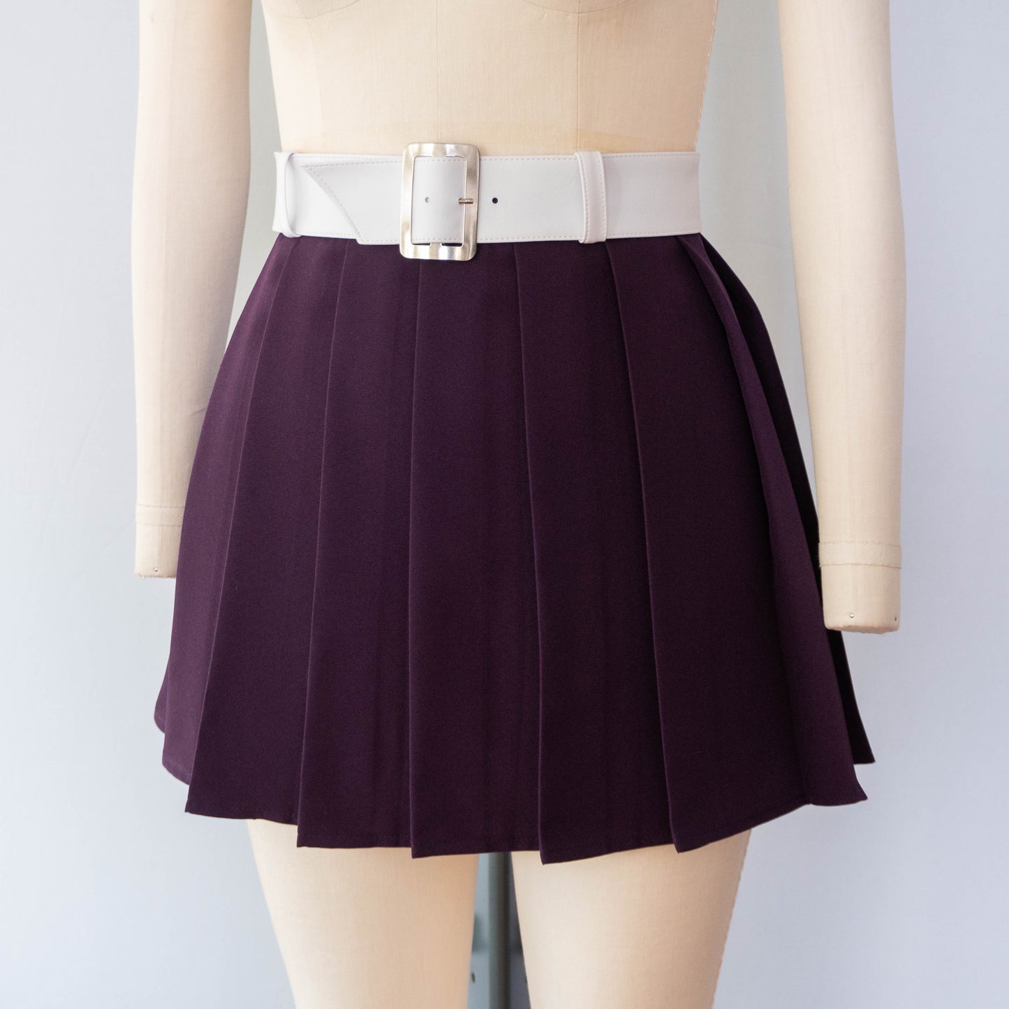 close-up studio shot of a dark purple pleated mini skirt with a thick white belt featuring a large silver rectangular buckle, shown on a mannequin.