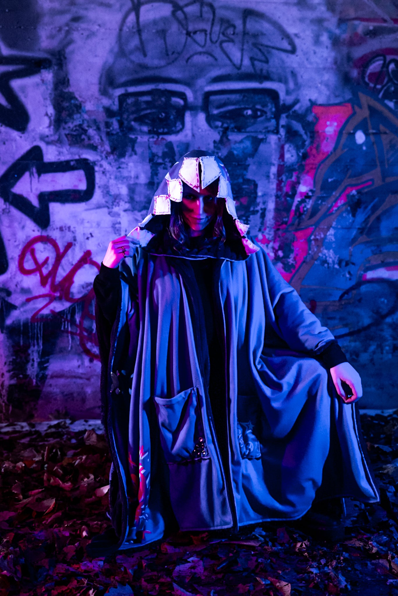 A cosplayer stands in an urban setting with graffiti-covered walls, illuminated by blue and purple lighting. The cosplayer is wearing a Arcane Jinx inspired purple blanket hoodie with large pockets adorned with whimsical patches and small studs. The hood shark theme and has patchwork shark teeth along the edges.