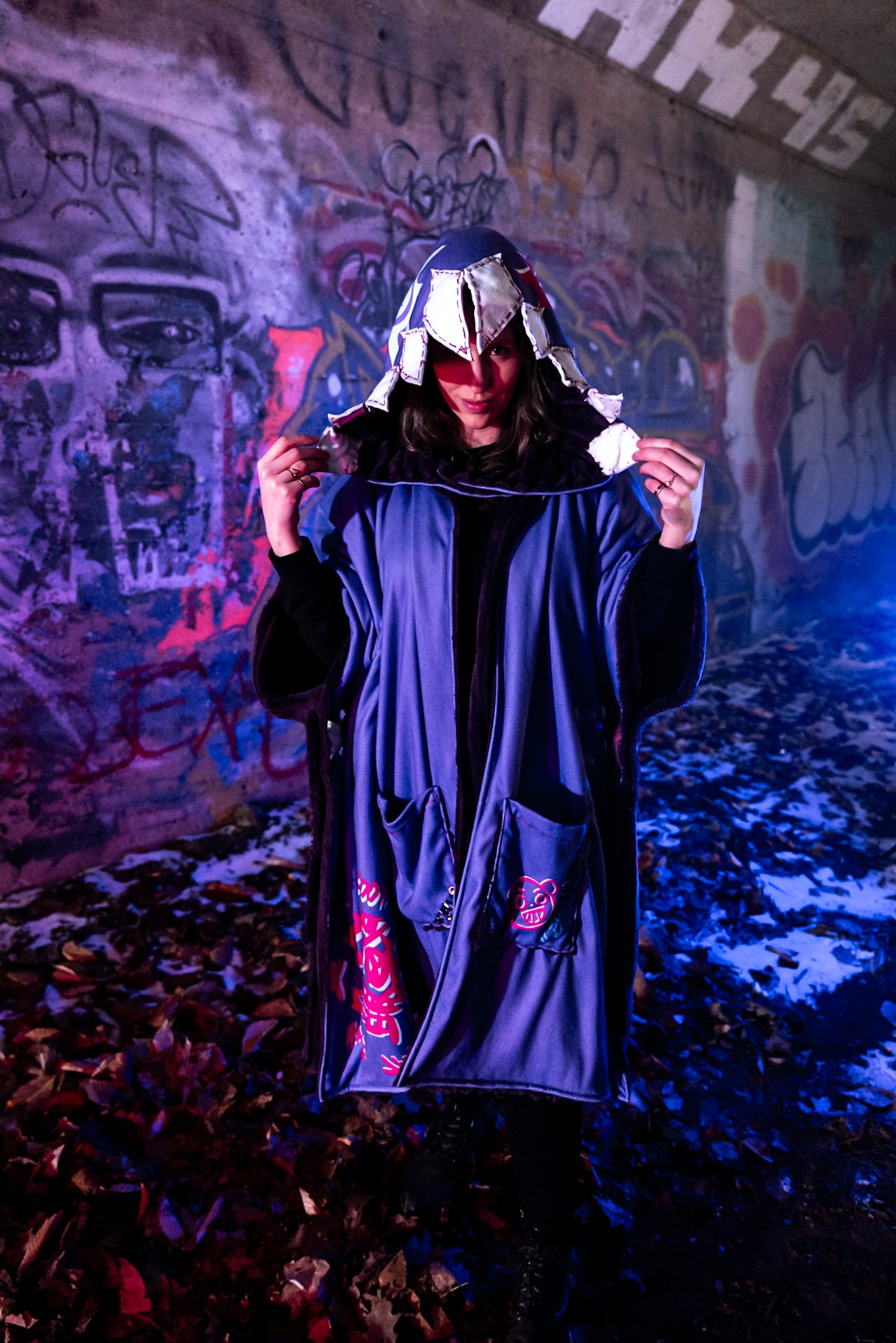 A cosplayer stands in an urban setting with graffiti-covered walls, illuminated by blue and purple lighting. The cosplayer is wearing a Arcane Jinx inspired purple blanket hoodie with large pockets adorned with whimsical patches and small studs. The hood shark theme and has patchwork shark teeth along the edges.