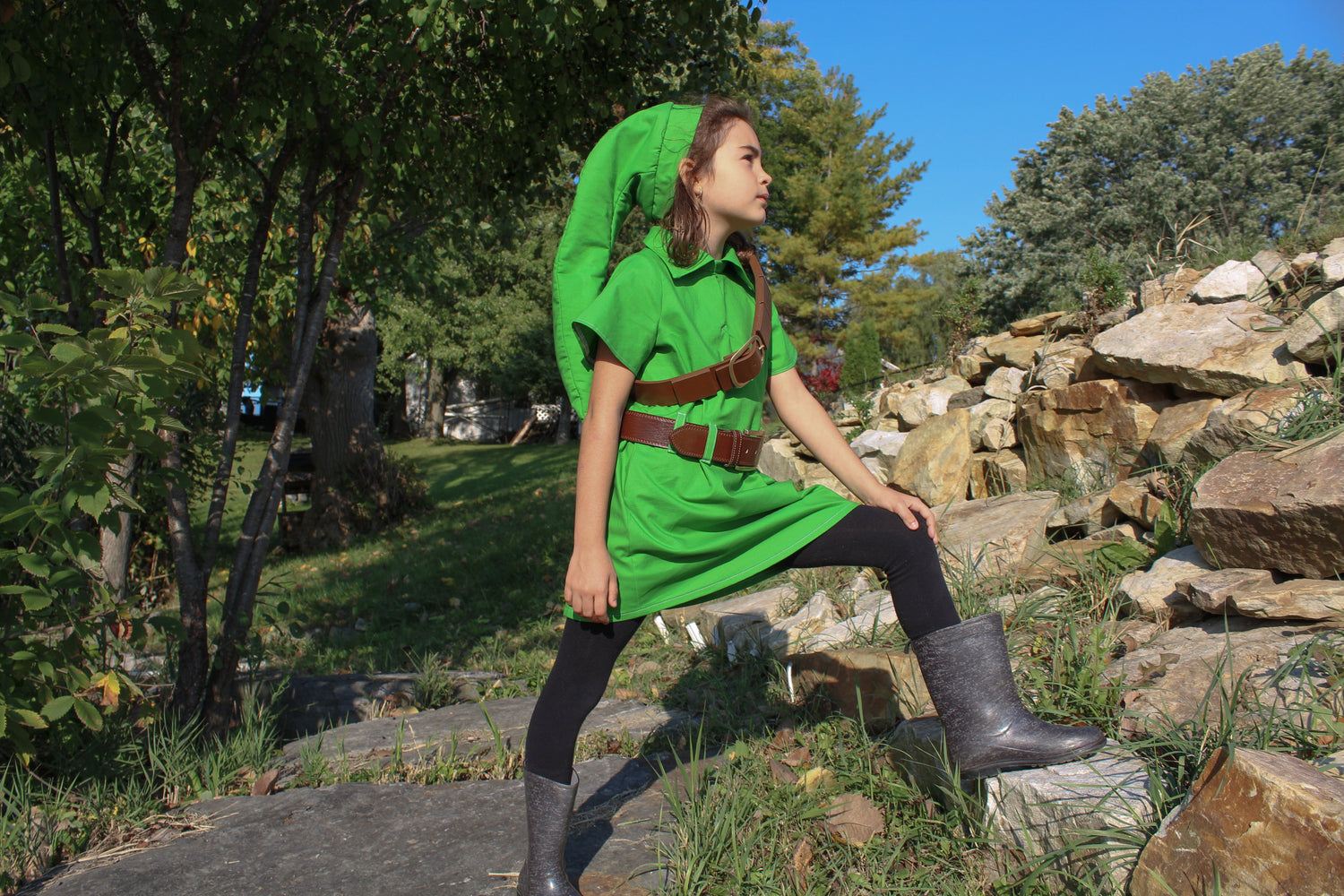 Young girl wearing a green cosplay tunic with a brown belt, black leggings, and boots, sitting on a rock in an outdoor park setting.