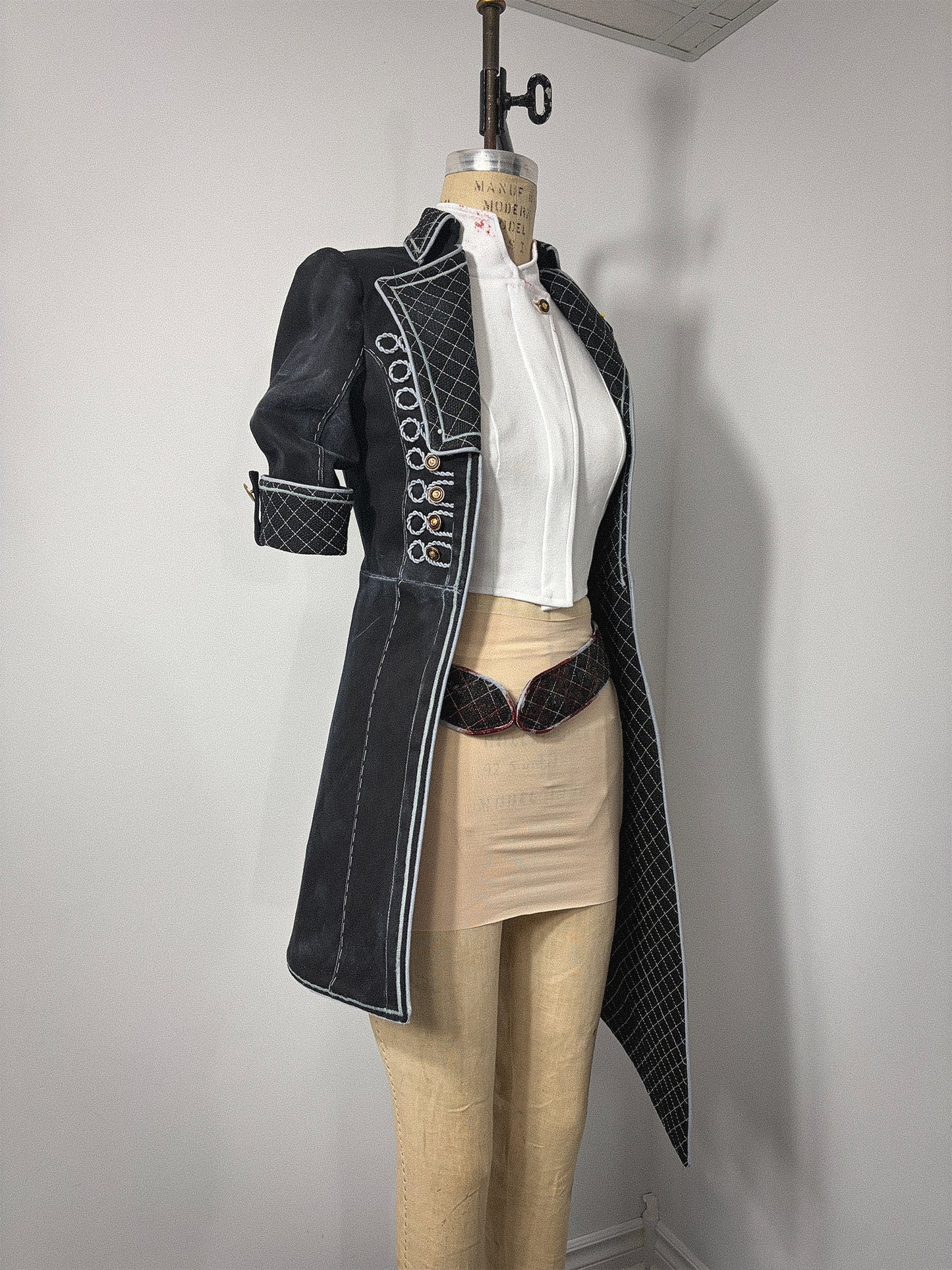 Side view of a cosplay jacket on a dress form, shown without the vest. Features a long black coat with white edge finishing, a vertical row of loop closures along the front opening, a short quilted sleeve, and a quilted inner lining visible at the hem. A white cropped top and dark quilted belt are visible underneath.
