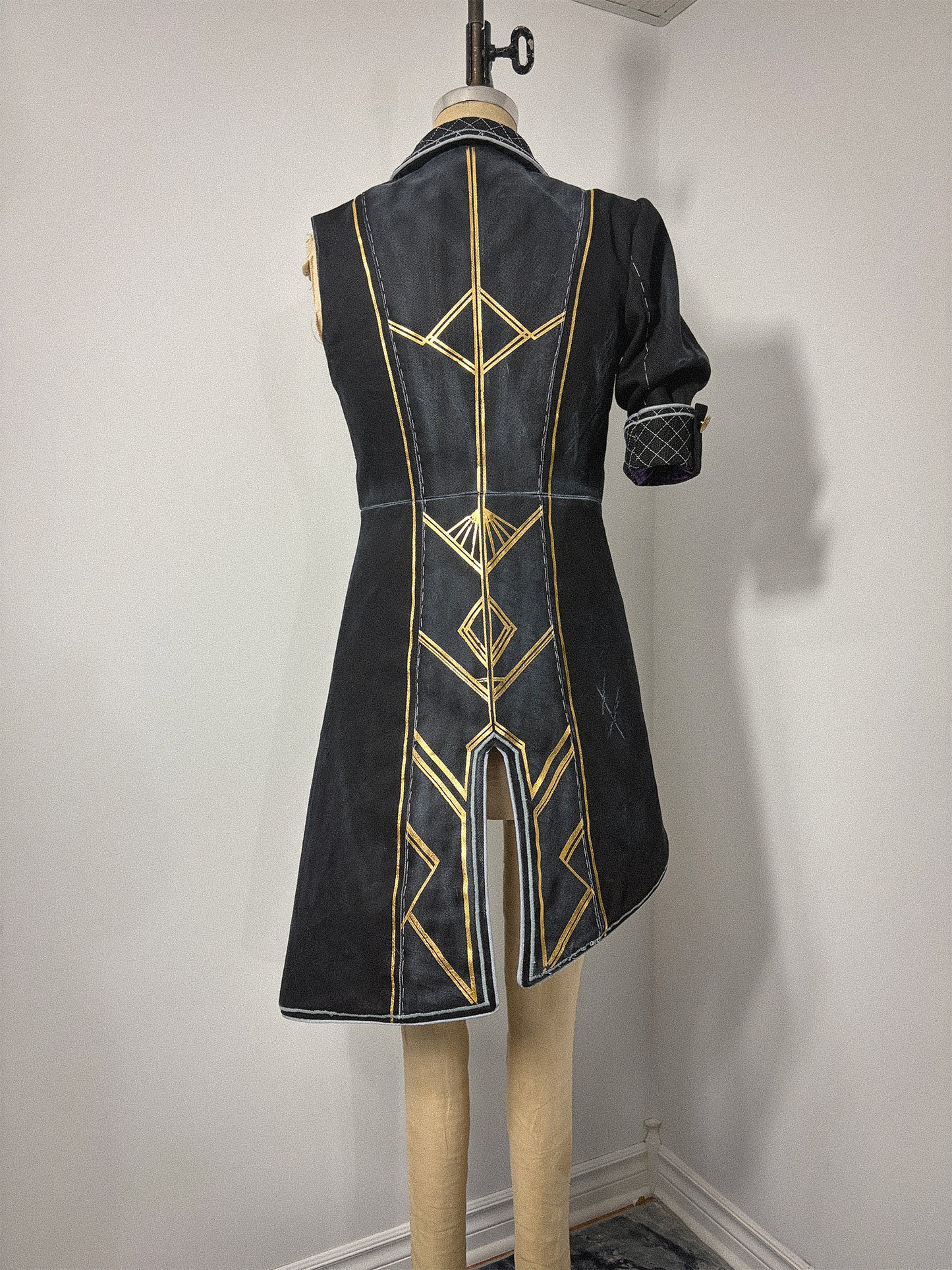 Back view of a completed Lune jacket cosplay from Clair Obscur: Expedition 33, displayed on a dress form. The black jacket features bold gold geometric designs across the back bodice and skirt panels, a center back vent, and a visible waist seam. The elbow-length quilted cuff is visible on the right sleeve. The asymmetrical hem sweeps longer on the left side.