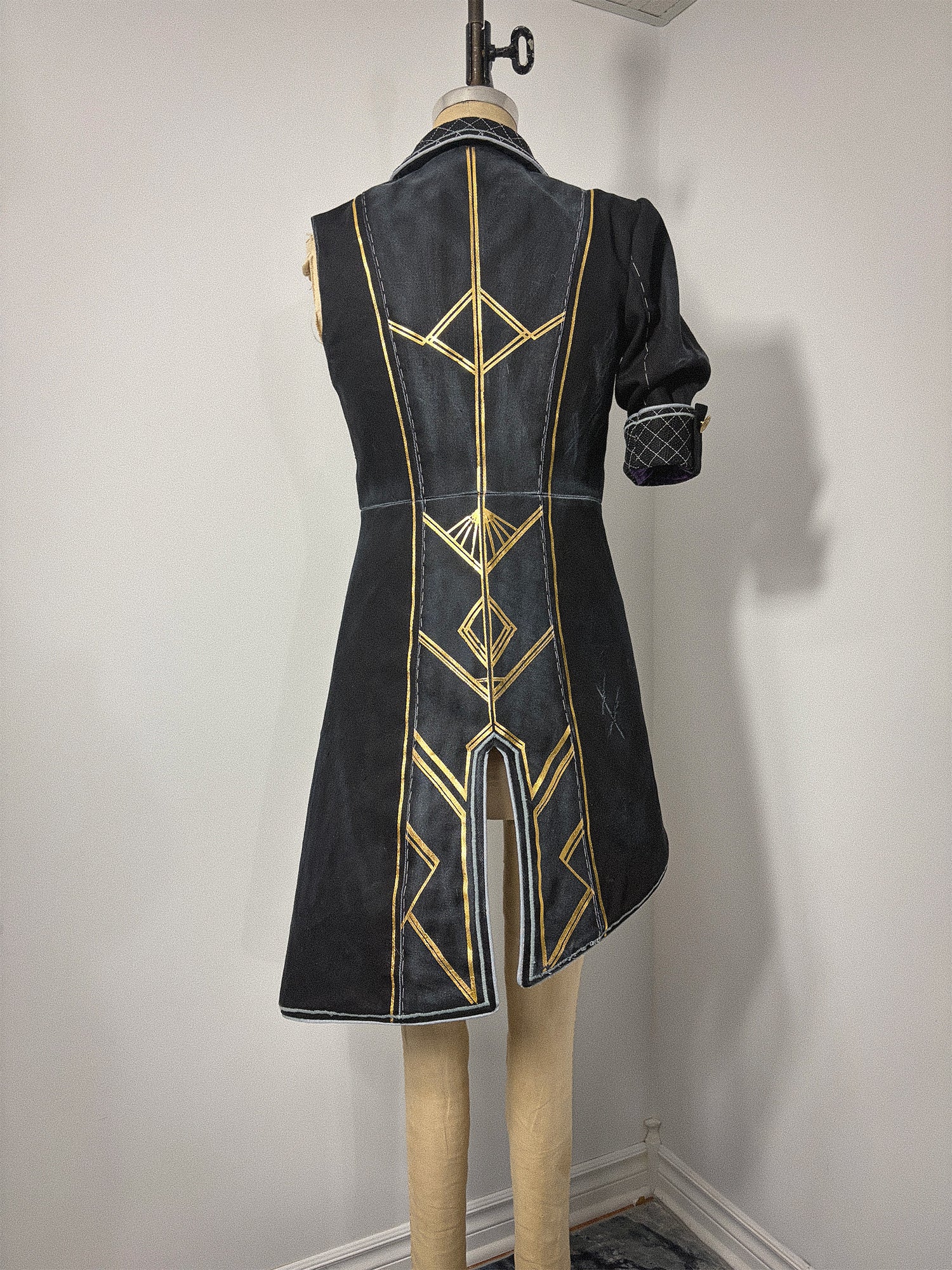 Back view of a completed Lune jacket cosplay from Clair Obscur: Expedition 33, displayed on a dress form. The black jacket features bold gold geometric designs across the back bodice and skirt panels, a center back vent, and a visible waist seam. The elbow-length quilted cuff is visible on the right sleeve. The asymmetrical hem sweeps longer on the left side.