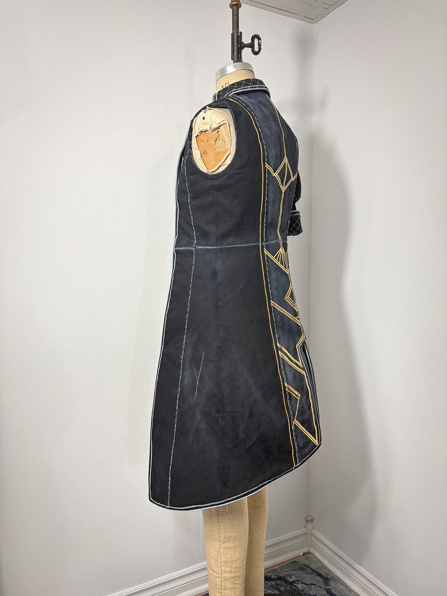 Side view of the Lune jacket cosplay from Clair Obscur: Expedition 33 on a dress form, showing the sleeveless left side with its open armhole, the waist seam, and the longer hem of the skirt. Gold geometric designs are visible along the back panel edge. The quilted cuff of the right sleeve is partially visible at the back.