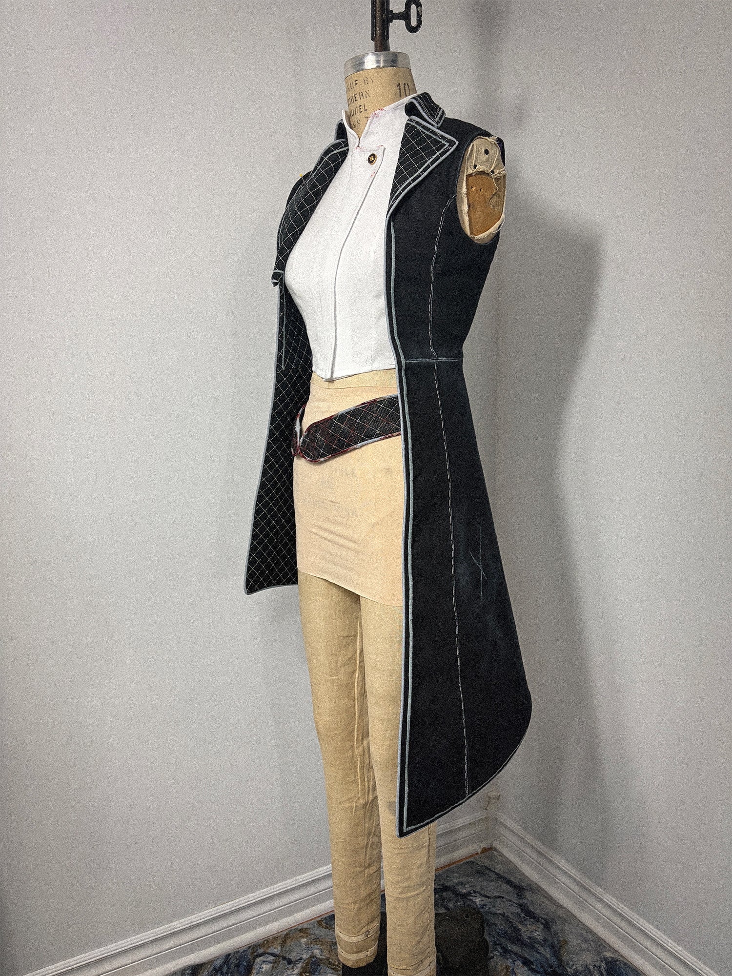 Side view of the Lune jacket cosplay from Clair Obscur: Expedition 33 on a full-length dress form, showing the sleeveless left side with its open armhole, quilted lapel, stand collar with gold button, and the long sweeping skirt hem reaching below the knee. A white undershirt and cream pants are visible beneath the open front jacket.