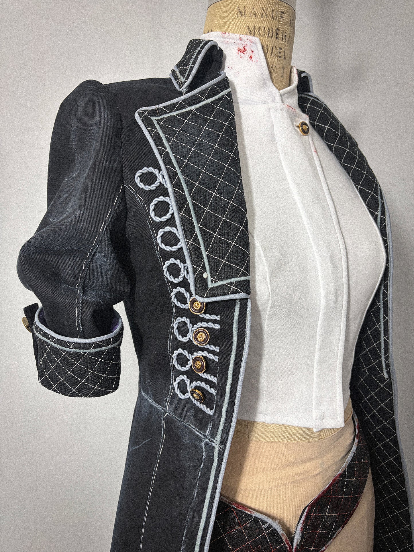 Close-up front view of the Lune jacket cosplay from Clair Obscur: Expedition 33 on a dress form, showing the right side bodice detail. Features decorative light grey corded button loops with gold buttons on the front panel, a quilted lapel, and an elbow-length sleeve with a quilted cuff and tab closure. A white undershirt is visible beneath the open front jacket.