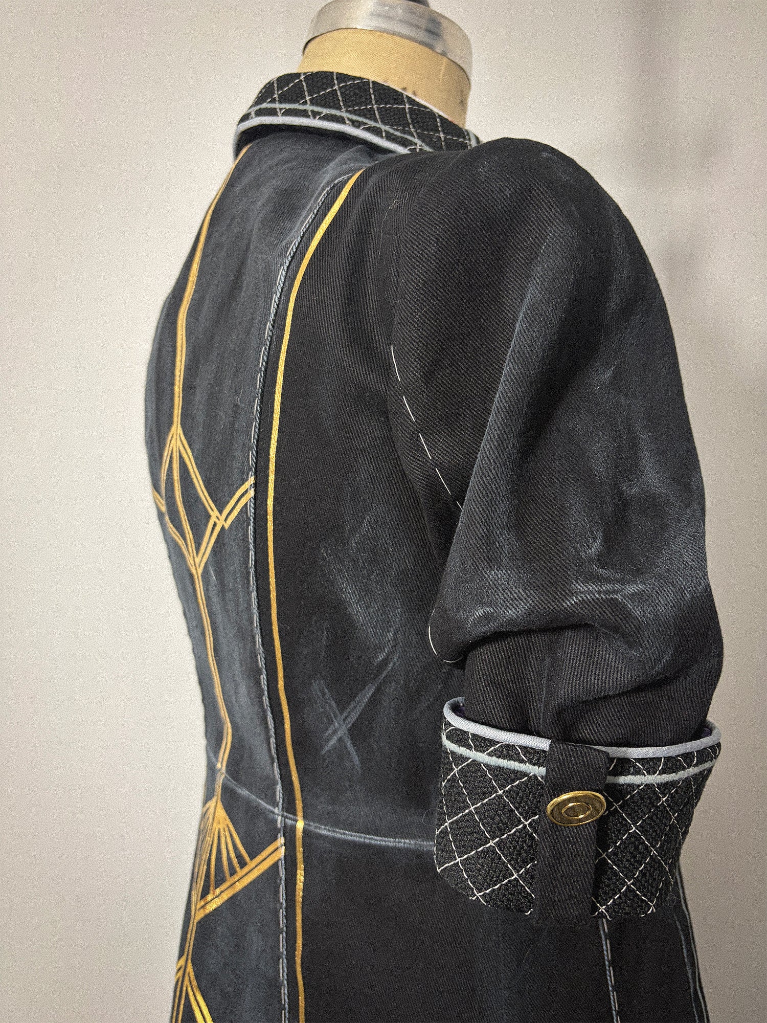 Close-up three-quarter back view of the Lune jacket cosplay from Clair Obscur: Expedition 33 on a dress form, highlighting the elbow-length sleeve and quilted cuff with its tab and gold button closure. Gold geometric designs and the princess seam are visible along the back bodice panel. The quilted collar is partially visible at the neckline.