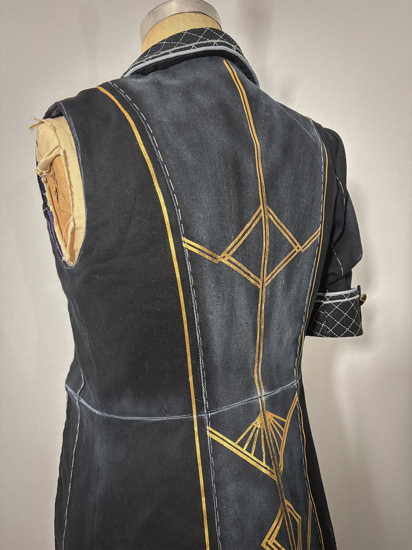 Three-quarter back view of the Lune jacket cosplay from Clair Obscur: Expedition 33 on a dress form, showing the sleeveless left side and the full back bodice and skirt with gold geometric designs spanning both panels across the waist seam. The princess seams, center back seam, and quilted collar are clearly visible. The sleeved right side with its quilted cuff and gold button is partially visible at the edge.