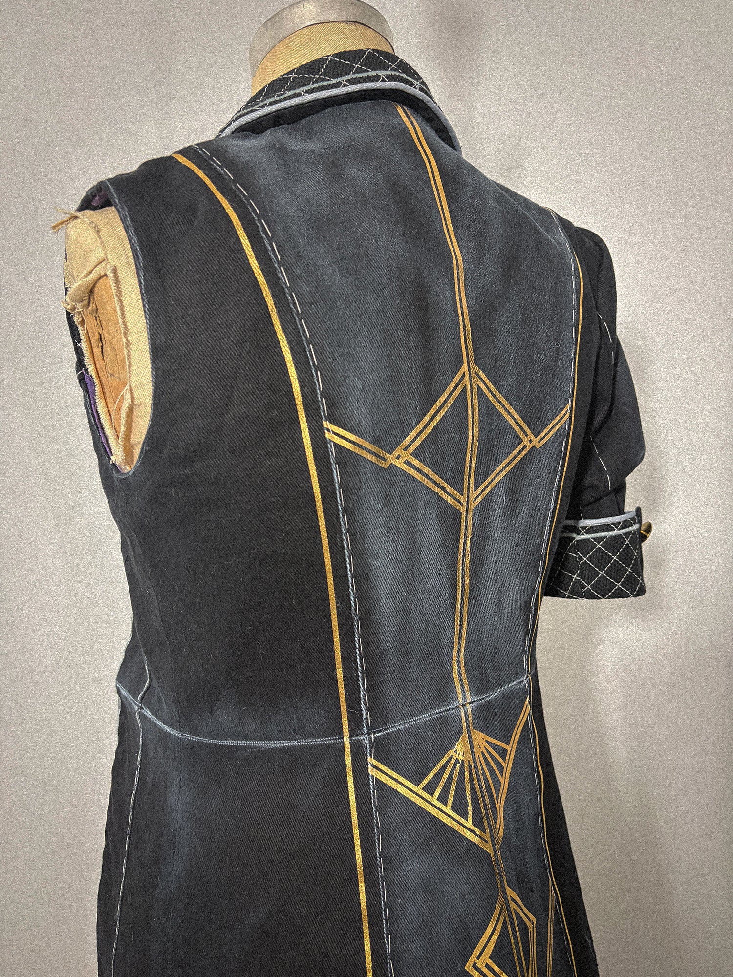 Three-quarter back view of the Lune jacket cosplay from Clair Obscur: Expedition 33 on a dress form, showing the sleeveless left side and the full back bodice and skirt with gold geometric designs spanning both panels across the waist seam. The princess seams, center back seam, and quilted collar are clearly visible. The sleeved right side with its quilted cuff and gold button is partially visible at the edge.