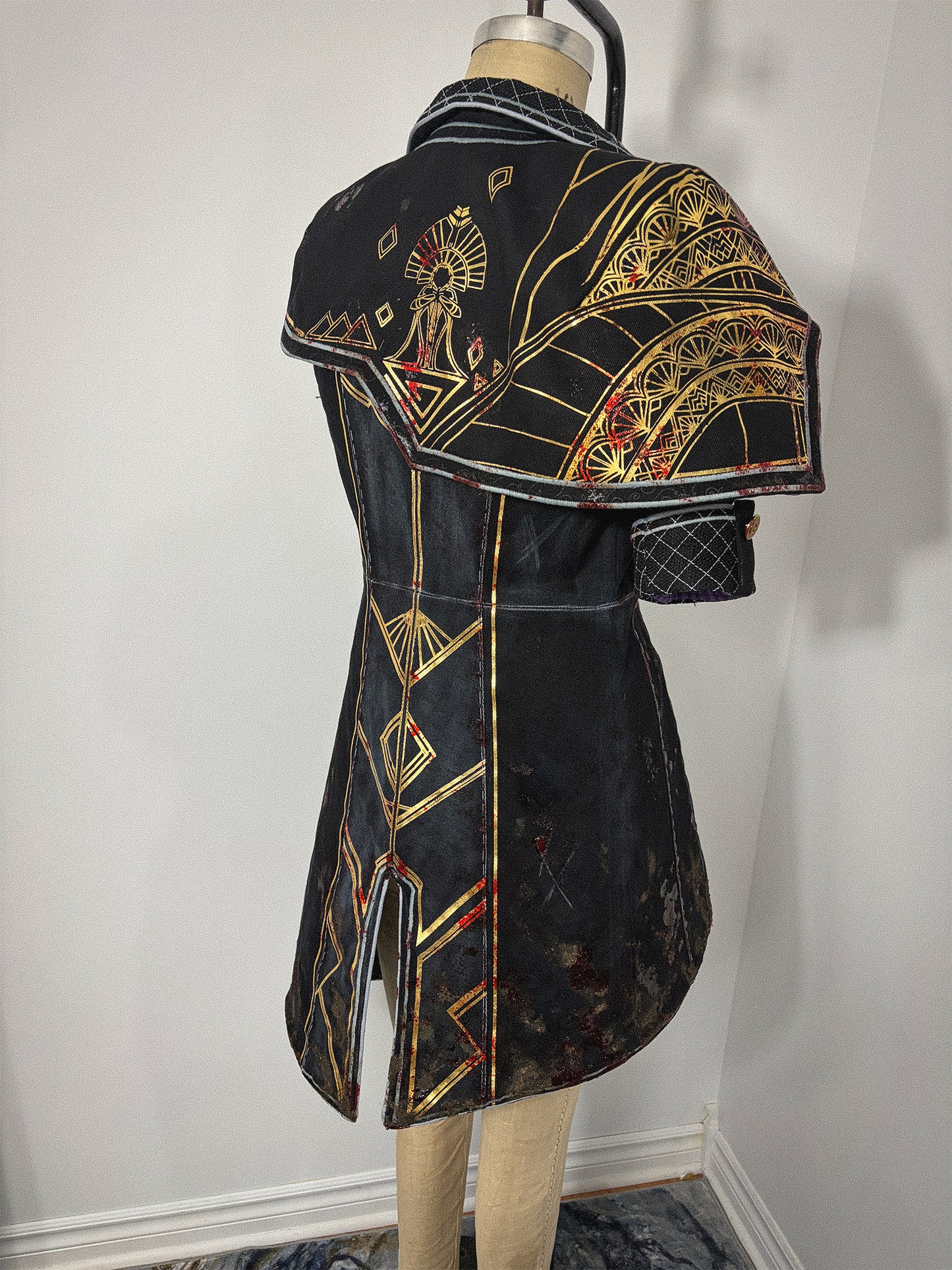 Three-quarter back view of a different Clair Obscur: Expedition 33 jacket cosplay on a dress form, featuring elaborate gold and red geometric and fan motif designs across the back bodice, skirt, and a structured capelet over the right shoulder. The center back vent, waist seam, princess seams, quilted collar, and quilted cuff are visible. The distressed fabric finish adds a battle-worn effect to the overall look.