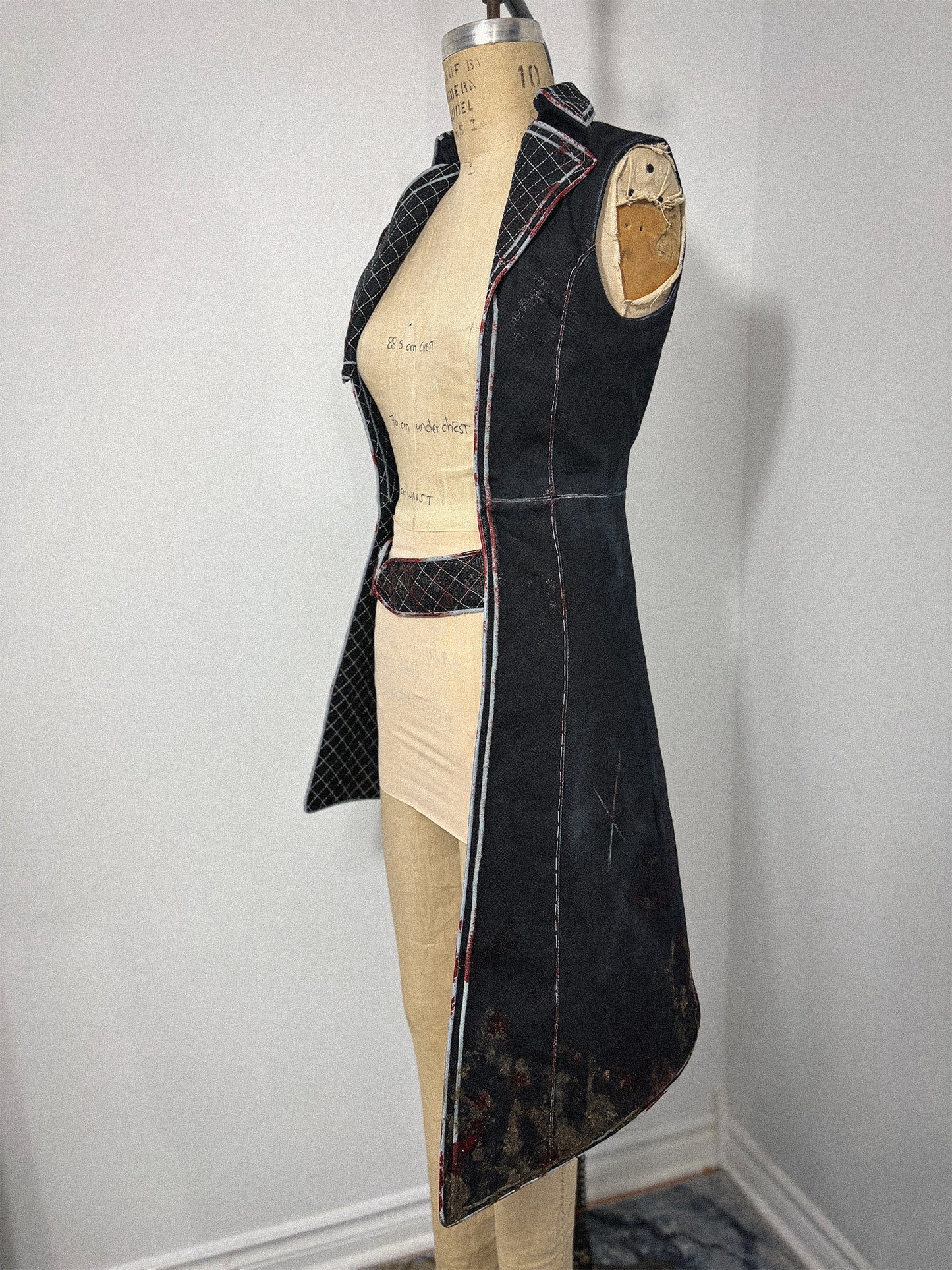 Side view of a Clair Obscur: Expedition 33 jacket cosplay variation on a full-length dress form, showing the sleeveless left side with its open armhole, quilted lapel, stand collar, and the long sweeping skirt hem with a distressed finish at the bottom edge. A quilted belt piece is visible at the waist. The open front reveals the bare dress form with handwritten measurements.