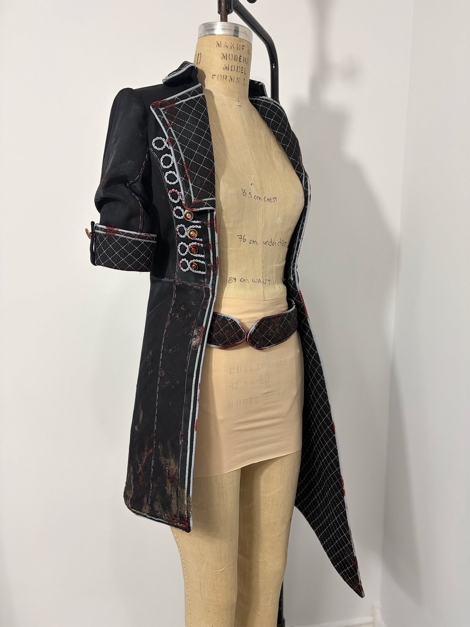Three-quarter front view of a Clair Obscur: Expedition 33 jacket cosplay variation on a dress form, showing the right side with corded button loops, quilted lapel, elbow-length sleeve with quilted cuff, and a distressed fabric finish with red accent details throughout. A quilted oval-buckle belt is visible at the waist. The open front reveals the bare dress form with handwritten measurements.