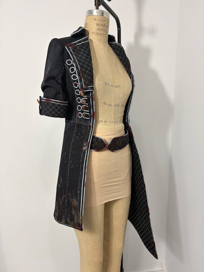 Three-quarter front view of a Clair Obscur: Expedition 33 jacket cosplay variation on a dress form, showing the right side with corded button loops, quilted lapel, elbow-length sleeve with quilted cuff, and a distressed fabric finish with red accent details throughout. A quilted oval-buckle belt is visible at the waist. The open front reveals the bare dress form with handwritten measurements.