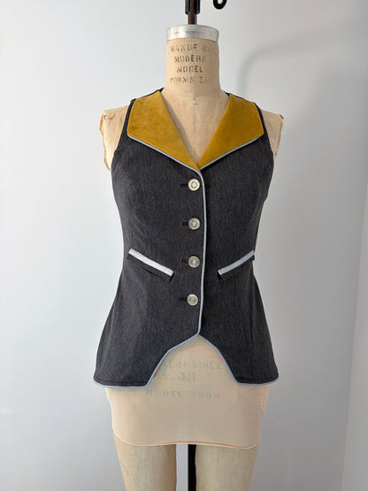 Front view of a fitted grey vest on a dress form. Features a deep V-neckline with gold lapels, four silver buttons down the center front, white piping trim along the lapels and pocket openings, and a curved pointed hem. Shown without the jacket or distressing.