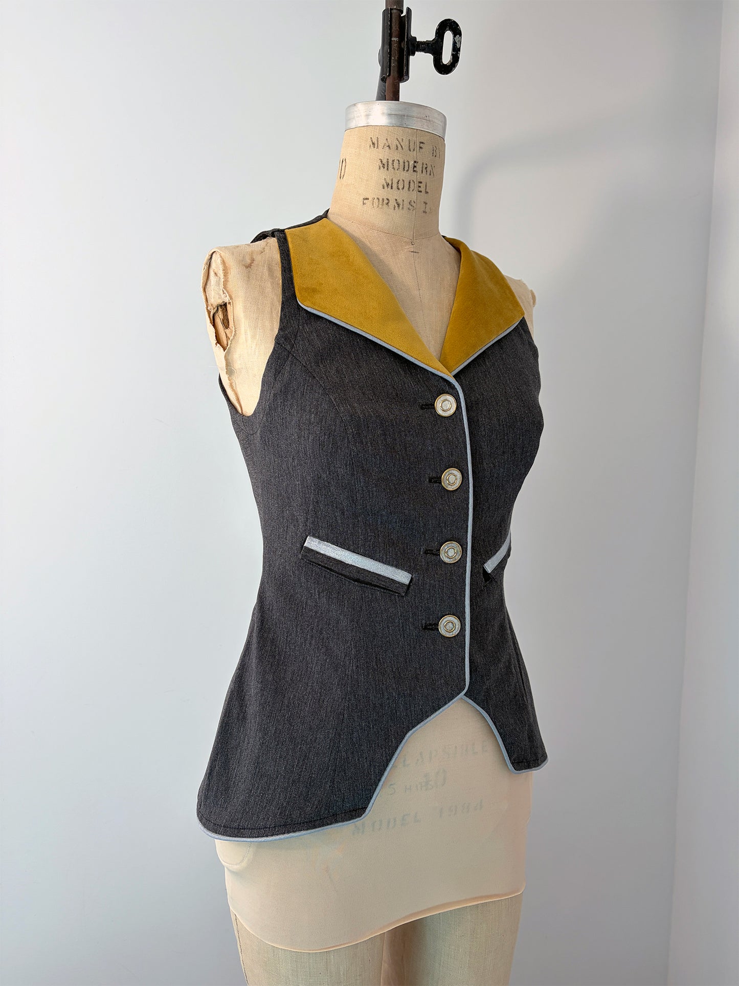 Three-quarter front view of the Structured Fitted Vest on a dress form, showing the full silhouette in dark grey fabric with a gold velvet lapel, light blue piping, welt pocket, four decorative buttons, and the asymmetrical pointed front hem.