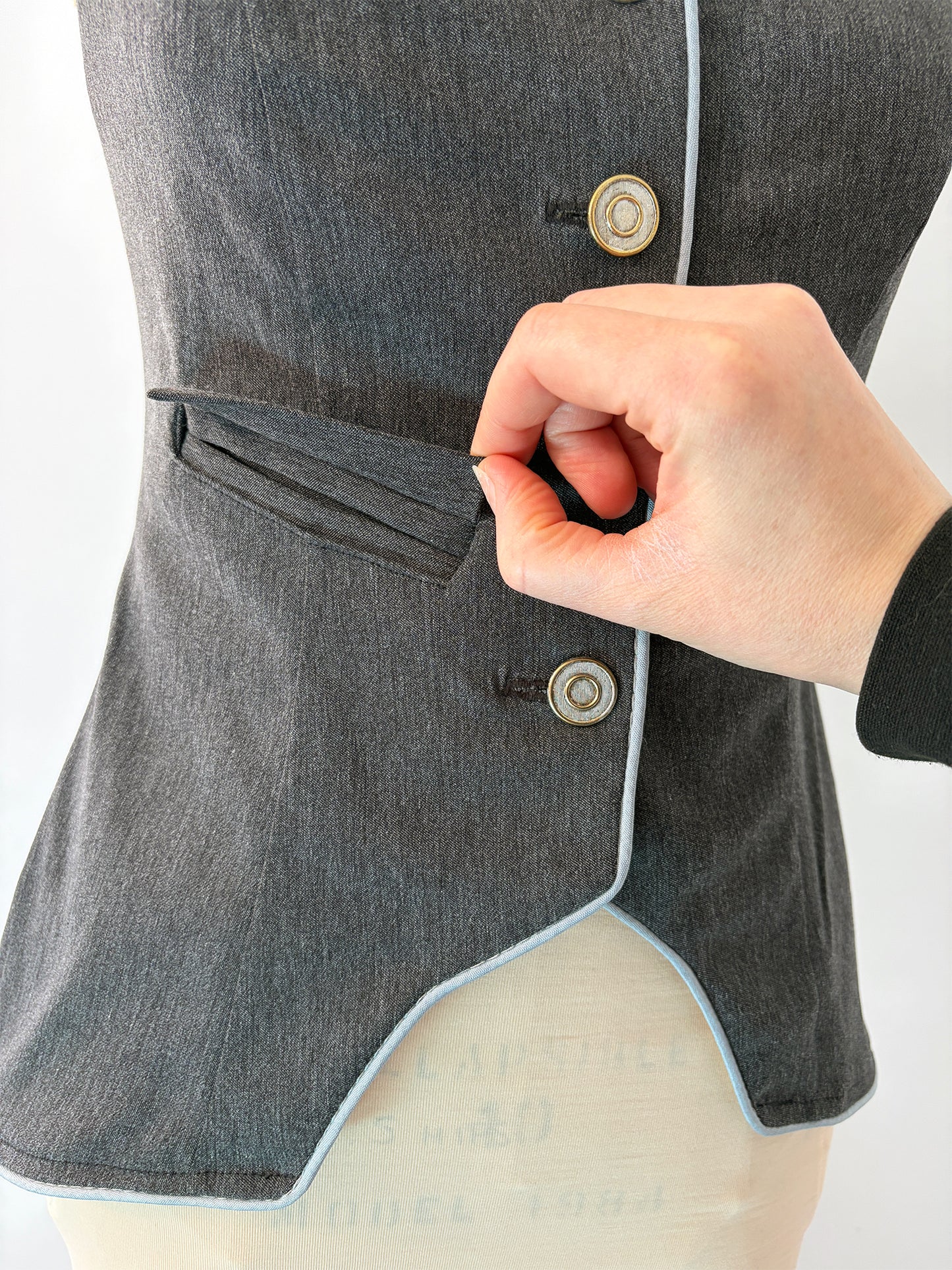 Close-up of a hand opening the welt pocket on the Structured Fitted Vest. The pocket sits cleanly above the waist on the grey fabric, flanked by two decorative buttons and finished with light blue piping along the curved hem edge.