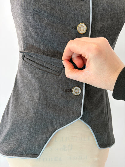 Close-up of a hand opening the welt pocket on the Structured Fitted Vest. The pocket sits cleanly above the waist on the grey fabric, flanked by two decorative buttons and finished with light blue piping along the curved hem edge.