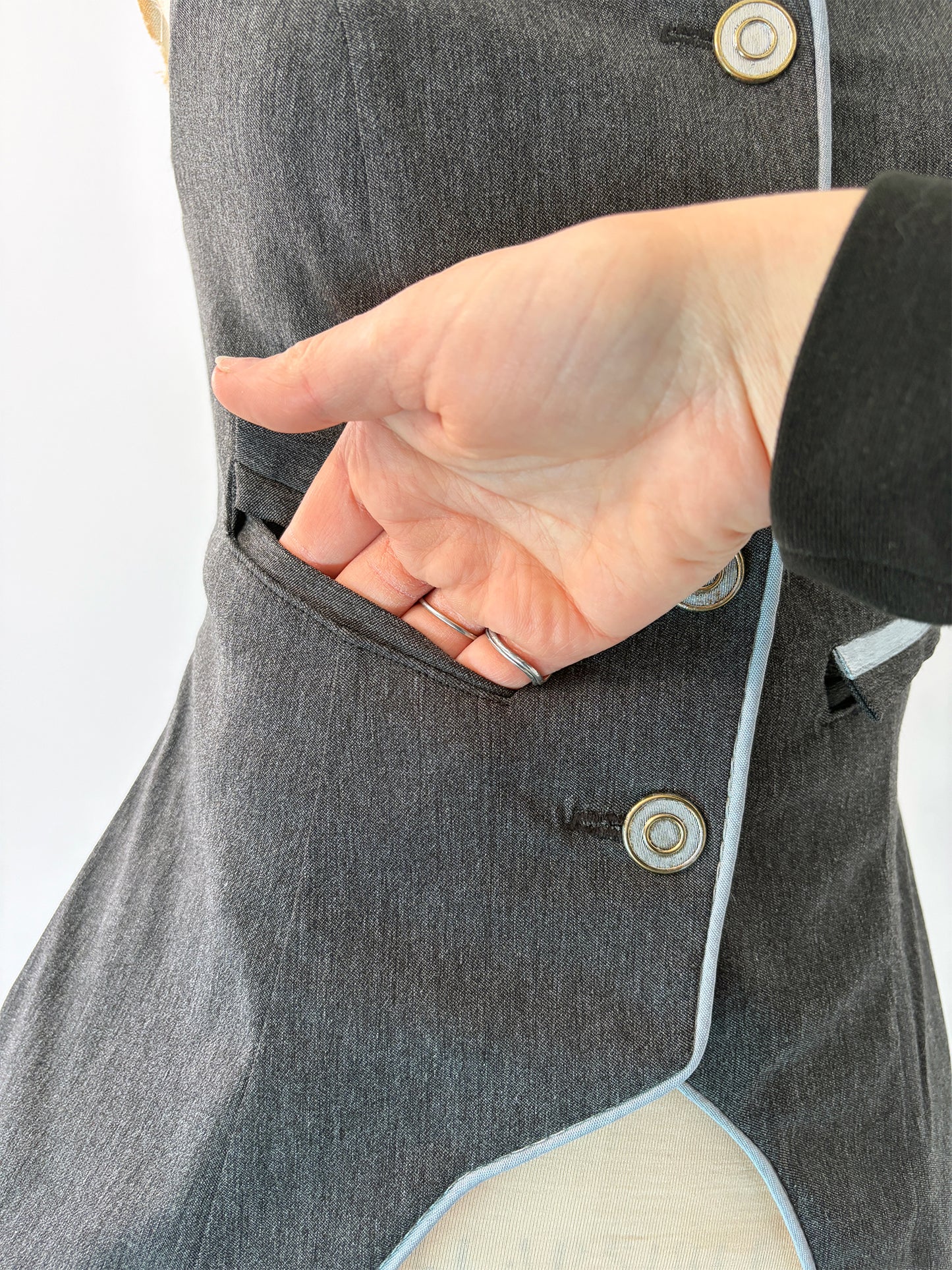 Close-up of a hand slipping inside the open welt pocket of the Structured Fitted Vest, showing the pocket depth and clean interior finish. Light blue piping trims the curved hem below.