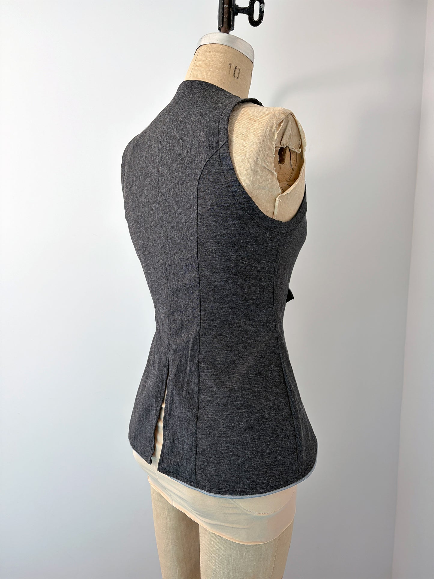 Back view of the Structured Fitted Vest on a dress form, in dark grey fabric with light blue piping along the hem. The back features princess seams for a close fit, a deep curved armhole, and a pointed hem that dips at center back.