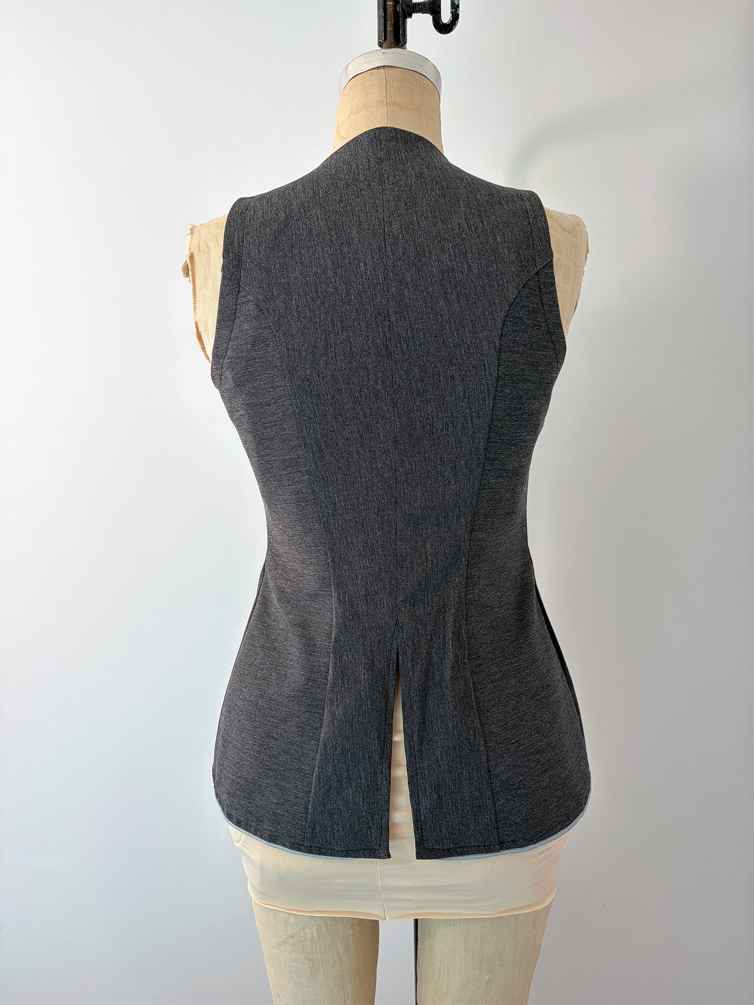 Back view of the Structured Fitted Vest on a dress form in dark grey fabric, showing the full back panel with princess seams, a defined waist, and a center back vent at the pointed hem.