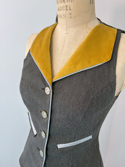 Close-up of the Structured Fitted Vest neckline on a dress form, highlighting the wide gold velvet lapel sewn into the shoulder seam, light blue piping along the front edge and pocket, and four decorative buttons down the center front.
