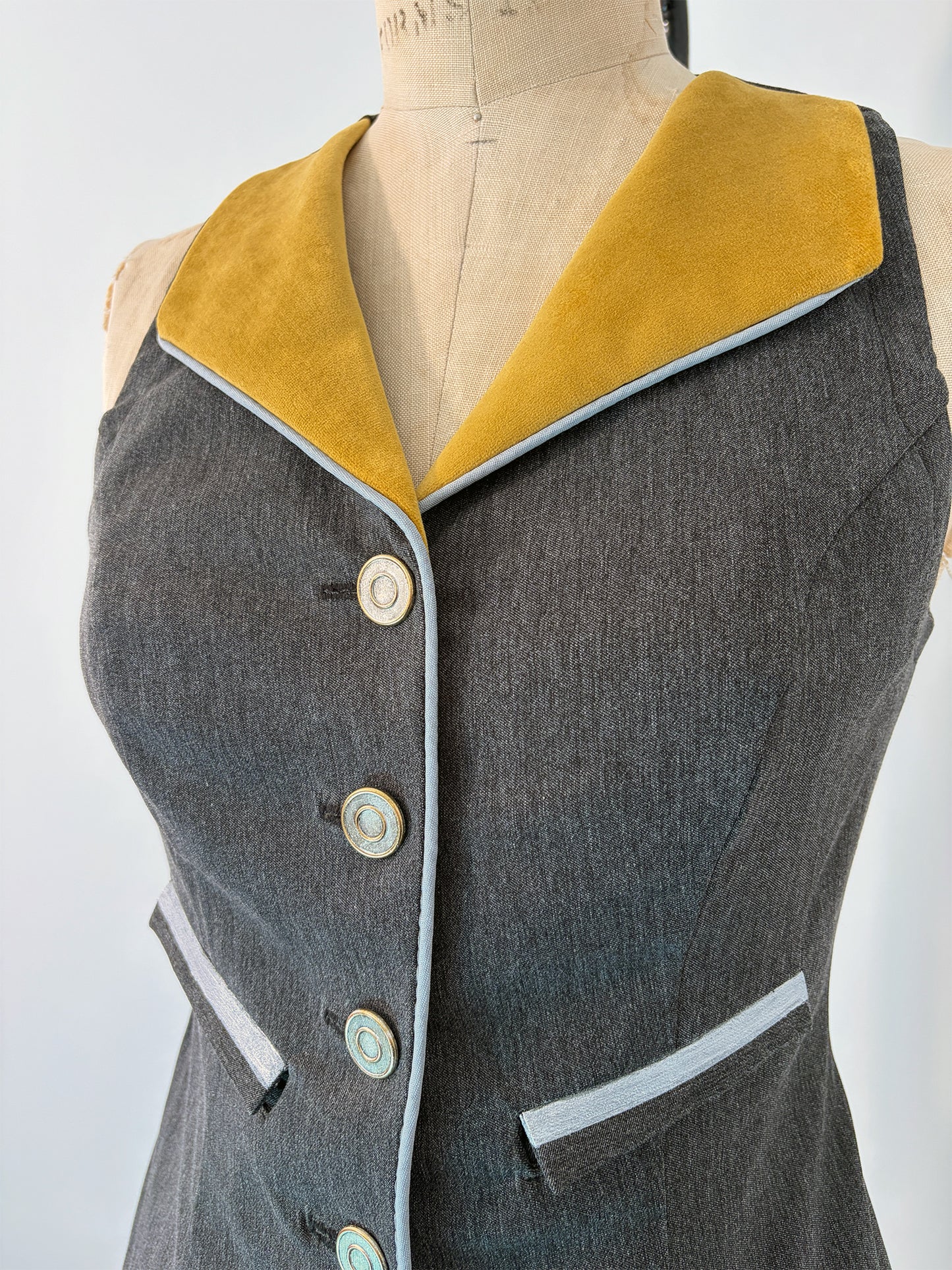 Front view of the Structured Fitted Vest on a dress form, showing the gold velvet lapel, five decorative buttons down the center front, light blue piping along the button band and welt pocket, and a fitted waist in dark grey fabric.