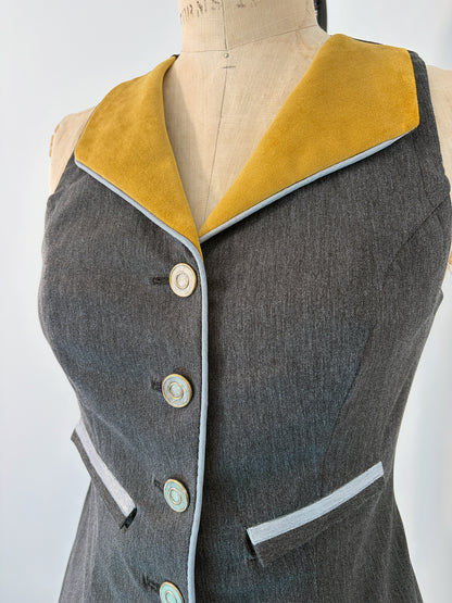 Front view of the Structured Fitted Vest on a dress form, showing the gold velvet lapel, five decorative buttons down the center front, light blue piping along the button band and welt pocket, and a fitted waist in dark grey fabric.