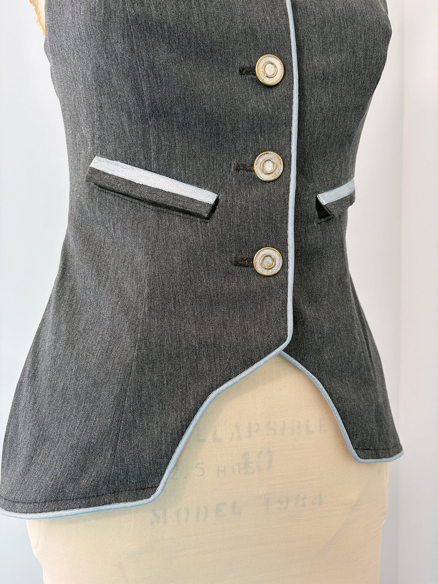 Close-up of the Structured Fitted Vest on a dress form, showing the front right panel in dark grey fabric with light blue piping along the hem and button placket. Three decorative buttons run down the center front. A welt pocket sits above the waist. The fitted silhouette curves into a pointed asymmetrical hem at the front.