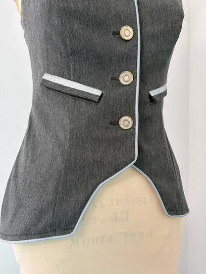 Close-up of the Structured Fitted Vest on a dress form, showing the front right panel in dark grey fabric with light blue piping along the hem and button placket. Three decorative buttons run down the center front. A welt pocket sits above the waist. The fitted silhouette curves into a pointed asymmetrical hem at the front.