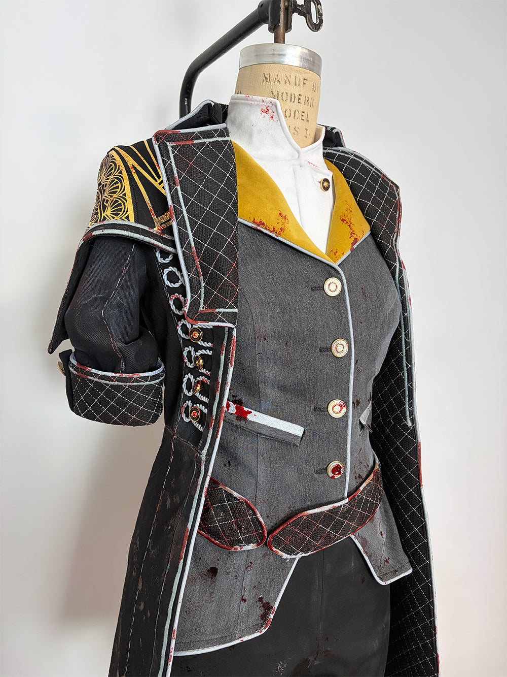 Close-up front detail of a cosplay costume on a dress form. Shows the grey four-button vest with a small chest pocket, gold and white layered lapels, and red-trimmed quilted black coat panels. The left shoulder piece features gold painted detailing, and the short quilted sleeve has embroidered or appliquéd lettering along the edge.