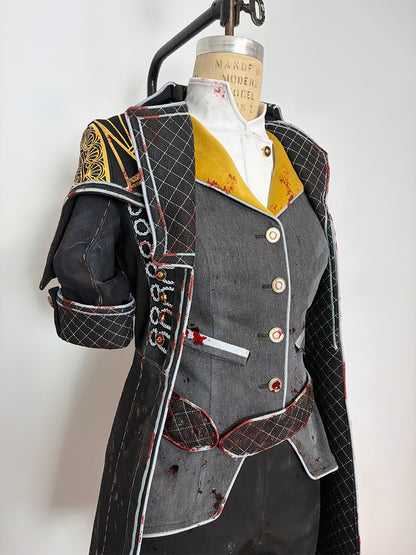 Close-up front detail of a cosplay costume on a dress form. Shows the grey four-button vest with a small chest pocket, gold and white layered lapels, and red-trimmed quilted black coat panels. The left shoulder piece features gold painted detailing, and the short quilted sleeve has embroidered or appliquéd lettering along the edge.