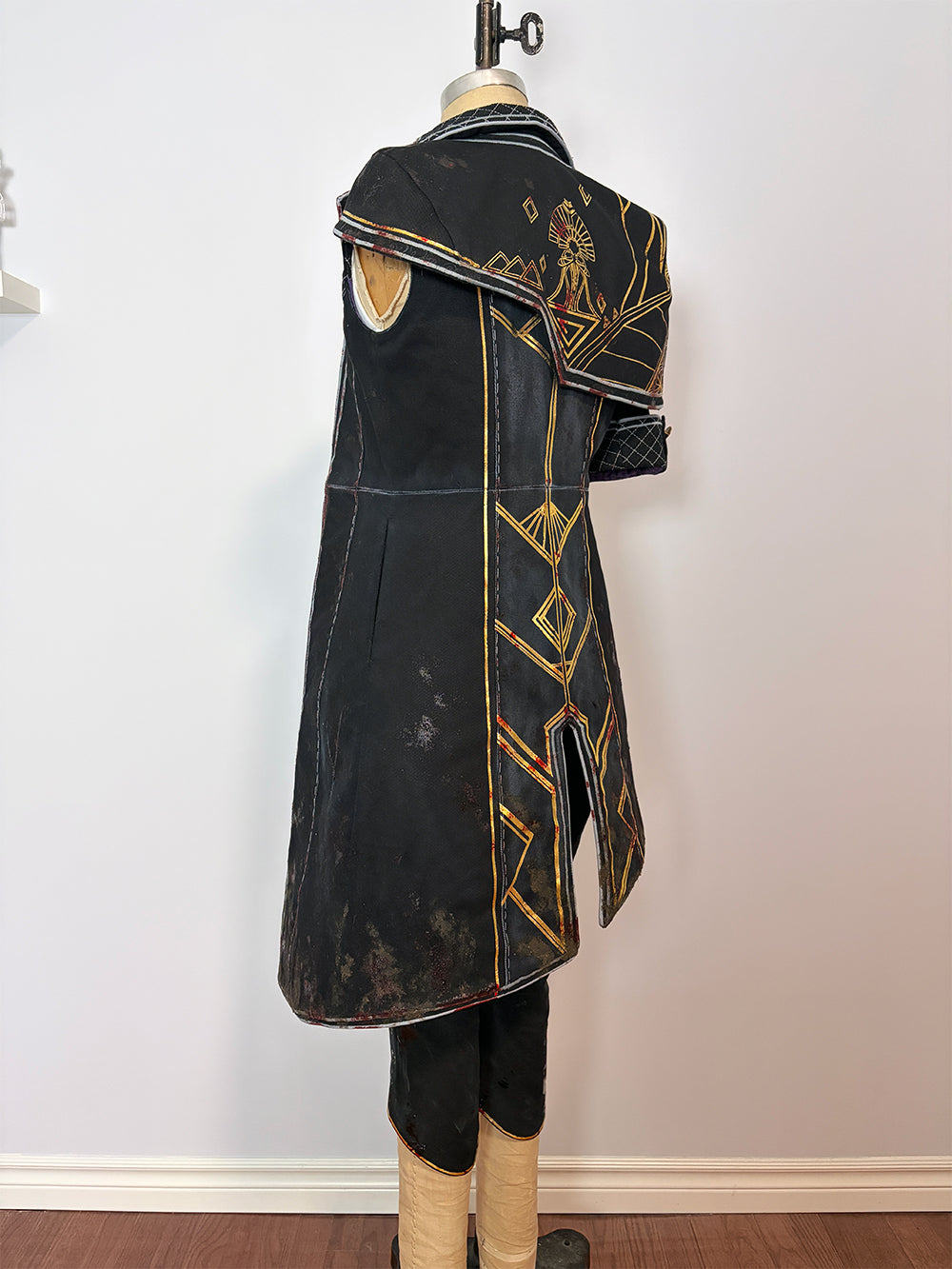 Back three-quarter view of a long black sleeveless cosplay coat on a dress form. Features gold and red geometric painted detailing across the back, a center back vent, visible lining at the hem, and a structured shoulder piece folded back to show the gold interior. Paired with black pants.