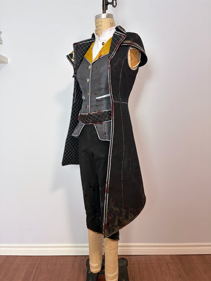 Side view of a cosplay costume on a dress form. Shows the asymmetrical hem of the long quilted black coat with red trim, a grey vest over a gold and white shirt, cropped black pants, and a structured shoulder piece. The coat's longer back panel and diagonal front opening are clearly visible from this angle.