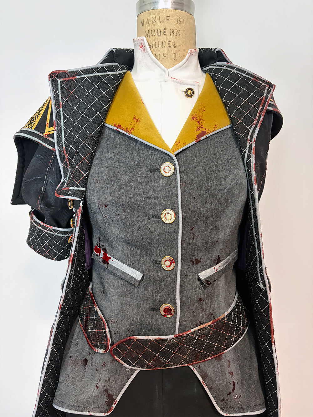 Front detail of a cosplay costume on a dress form. Features a grey four-button vest with welt pockets and blood splatter distressing, wide gold lapels, a white button-up underneath, and red-trimmed quilted black coat panels on either side. The structured shoulder piece and short quilted sleeve are visible on the left.