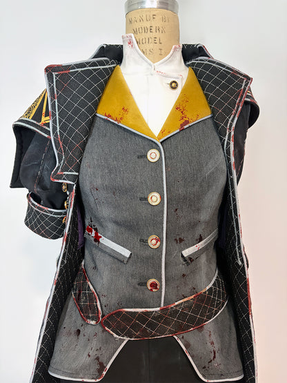 Front detail of a cosplay costume on a dress form. Features a grey four-button vest with welt pockets and blood splatter distressing, wide gold lapels, a white button-up underneath, and red-trimmed quilted black coat panels on either side. The structured shoulder piece and short quilted sleeve are visible on the left.