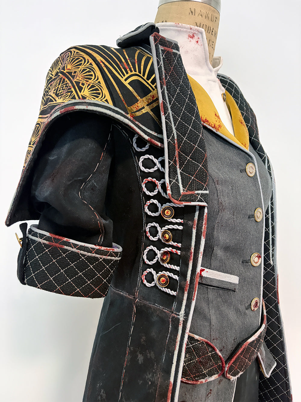 Side detail of a cosplay costume on a dress form. Shows the structured quilted shoulder piece with gold painted fan motifs, the short quilted sleeve with a vertical row of loop closures, red-trimmed coat panels, and the side of the grey vest with button detail and a metal bar embellishment. Gold lapel and white undershirt are visible at the neckline.