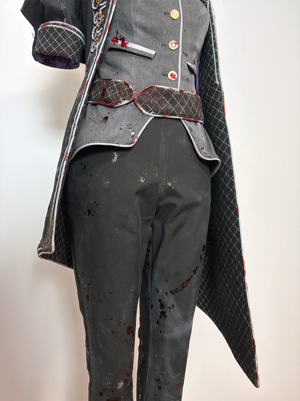Lower body detail of a cosplay costume on a dress form. Shows the asymmetrical hem of the quilted black coat with red trim, the curved bottom edge of the grey vest, a quilted dark red belt with scalloped edges, black cropped pants with blood splatter distressing, and the short quilted sleeve with a purple lining visible at the cuff.