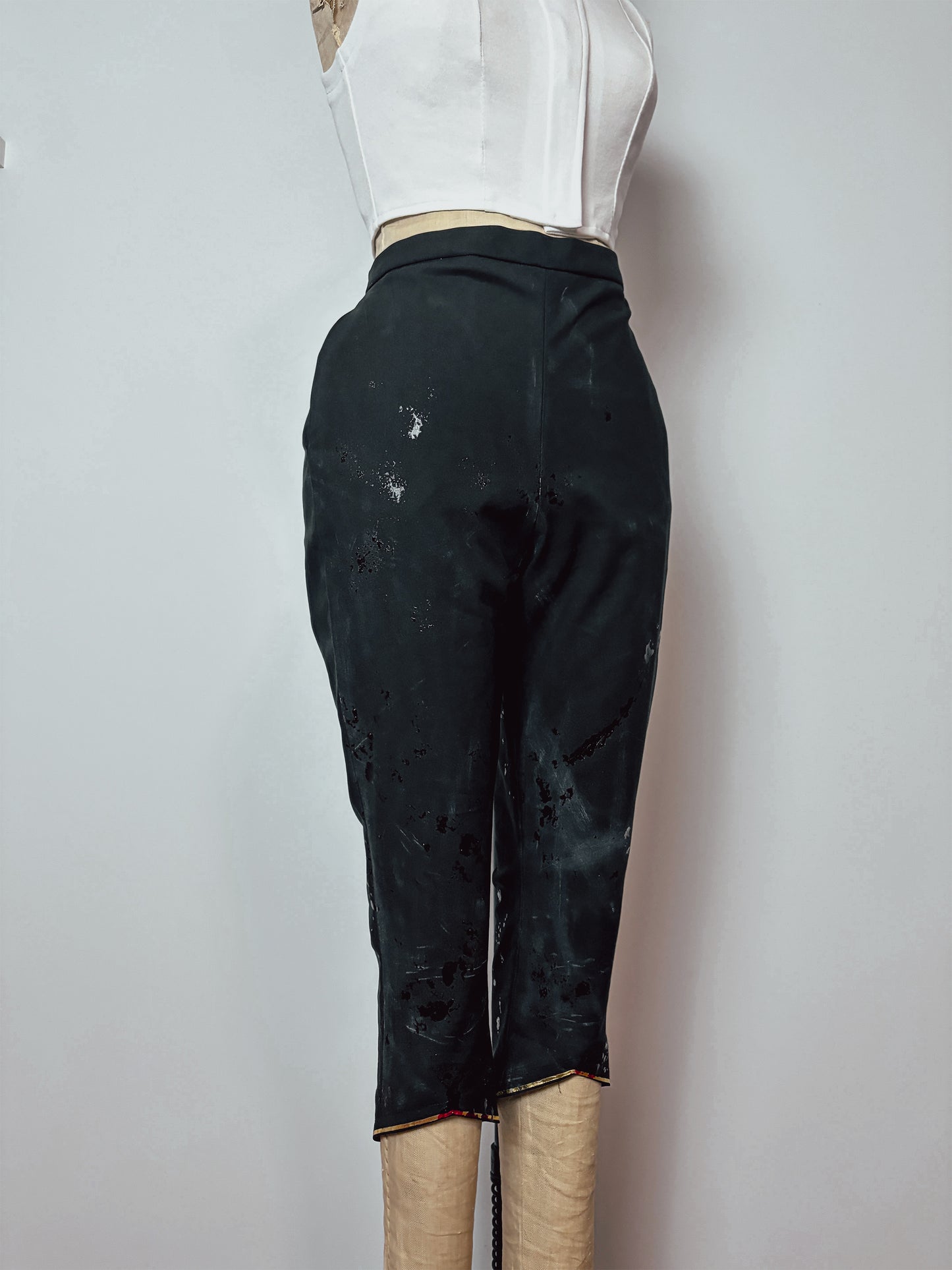 Front view of cropped black cosplay pants on a dress form. Features a high waist, slim fit, and heavy blood splatter distressing across the front. A subtle gold or orange trim is visible at the cropped hem.