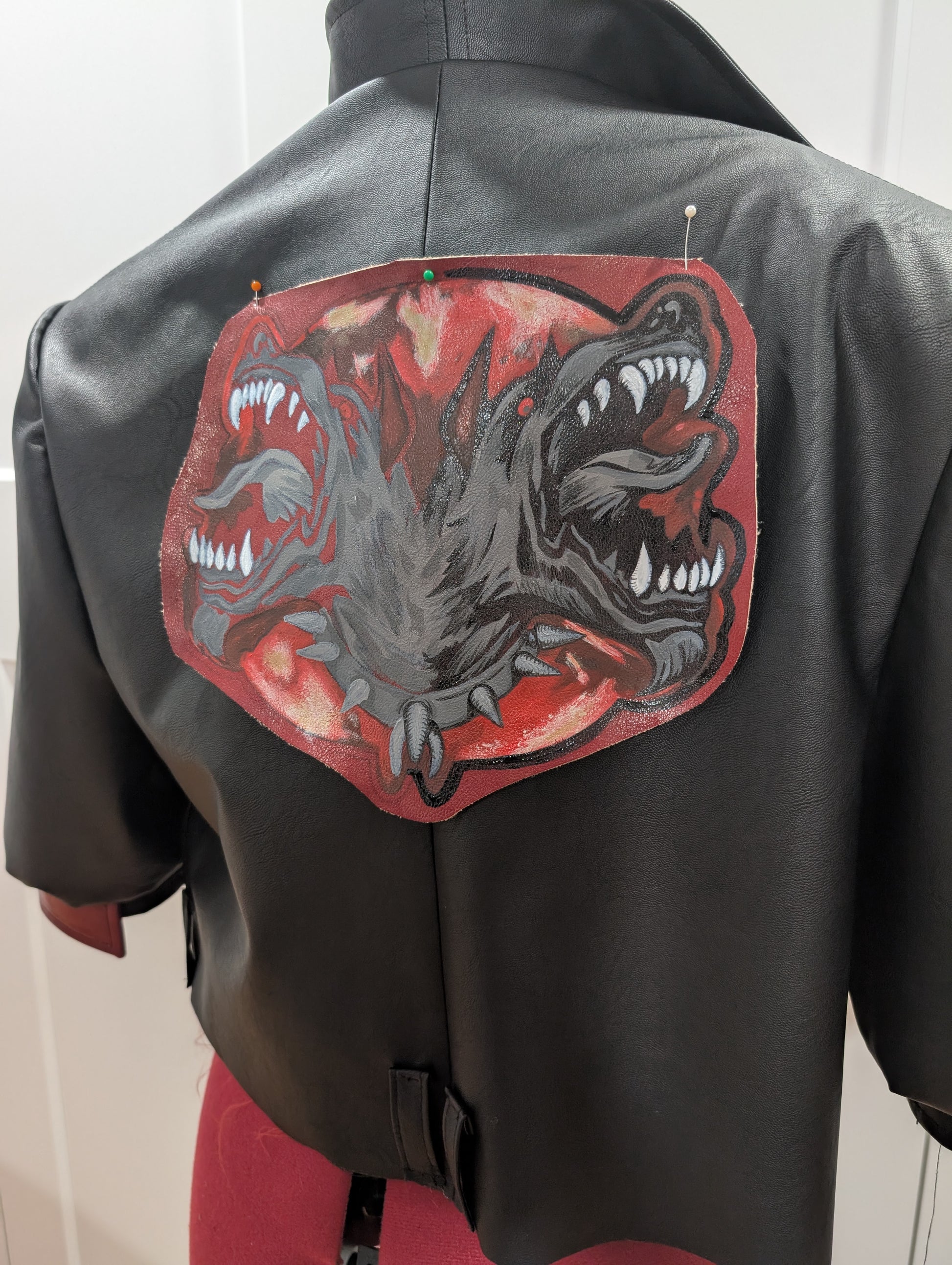 A black cropped jacket with a hand-painted patch of a snarling two-headed dog, pinned to the back for placement. The patch features red, black, and gray tones with sharp teeth and spiked details.
