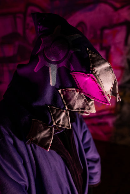 A cosplayer stands in an urban setting with graffiti-covered walls, illuminated by blue and purple lighting. The cosplayer is wearing a Arcane Jinx inspired purple blanket hoodie with large pockets adorned with whimsical patches and small studs. The hood shark theme and has patchwork shark teeth along the edges.