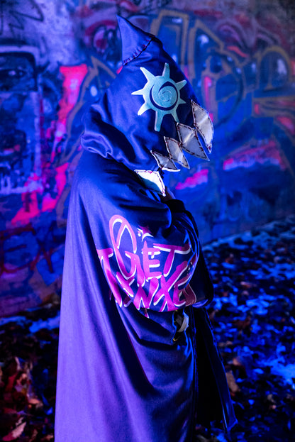 A cosplayer stands in an urban setting with graffiti-covered walls, illuminated by blue and purple lighting. The cosplayer is wearing a Arcane Jinx inspired purple blanket hoodie with large pockets adorned with whimsical patches and small studs. The hood shark theme and has patchwork shark teeth along the edges.