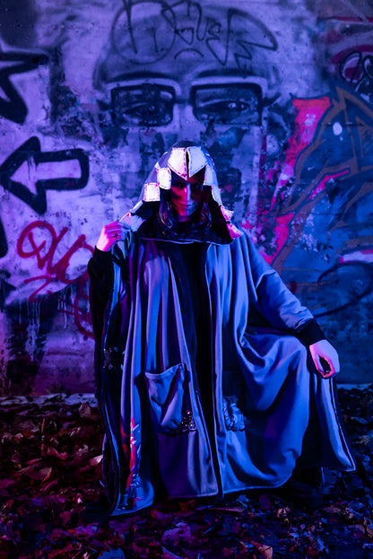 A cosplayer stands in an urban setting with graffiti-covered walls, illuminated by blue and purple lighting. The cosplayer is wearing a Arcane Jinx inspired purple blanket hoodie with large pockets adorned with whimsical patches and small studs. The hood shark theme and has patchwork shark teeth along the edges.