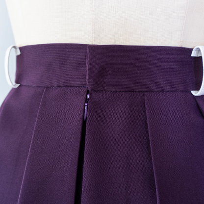 close-up studio shot of a dark purple pleated mini skirt showing an invisible zipper at the center back, shown on a mannequin.