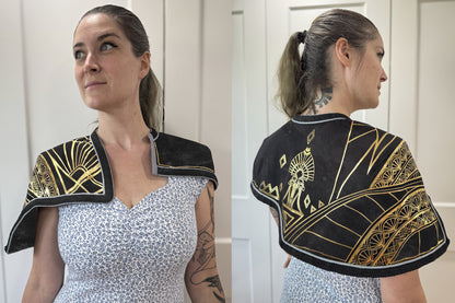 Front and back views of a black capelet worn by a person over a floral dress. The capelet features extensive gold geometric and Art Deco-style painted or HTV detailing, including fan motifs, diamond shapes, and a central figure on the back. The edges are finished with a fine red trim.